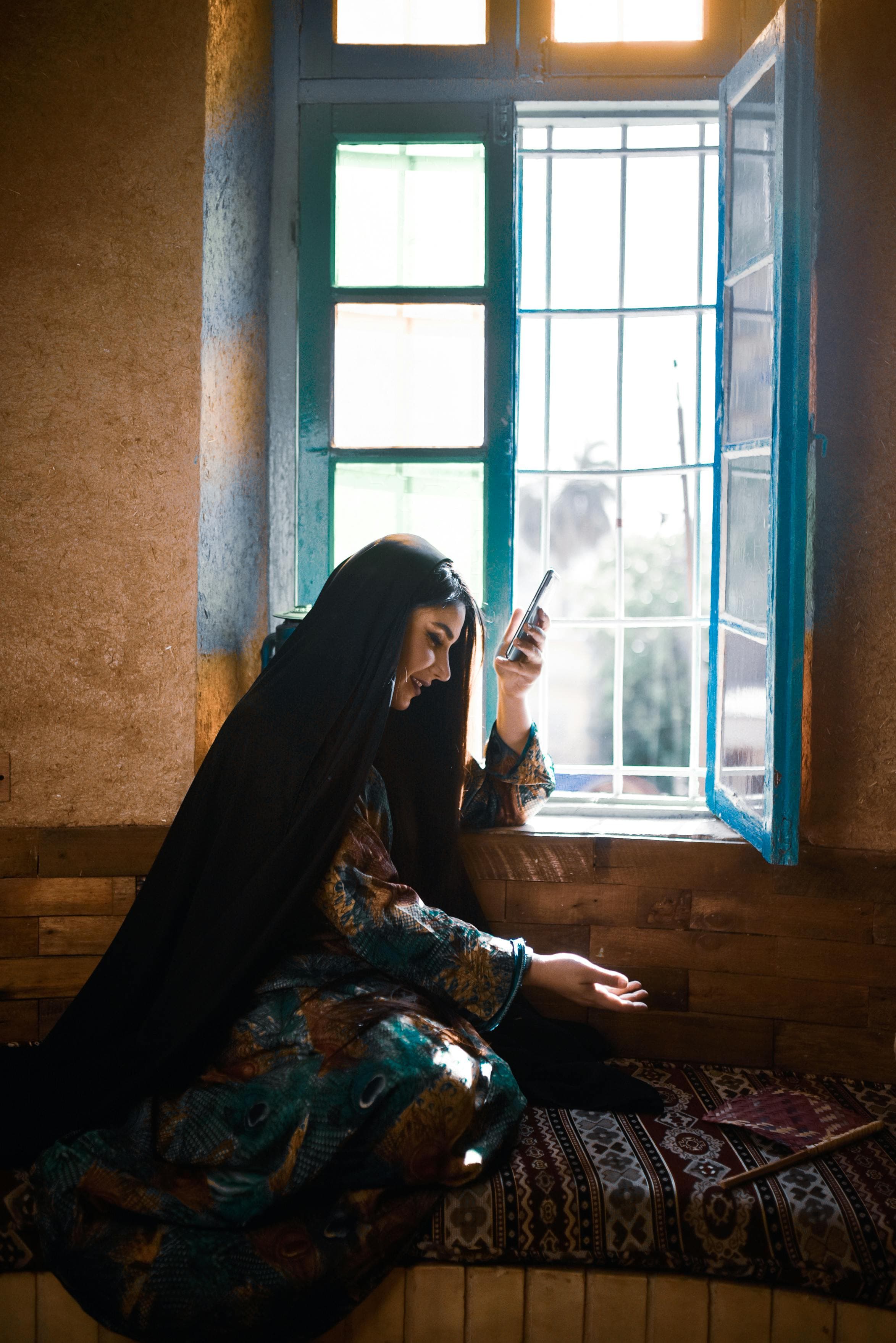 Femme afghane assise près d'une fenêtre bleue tenant son téléphone