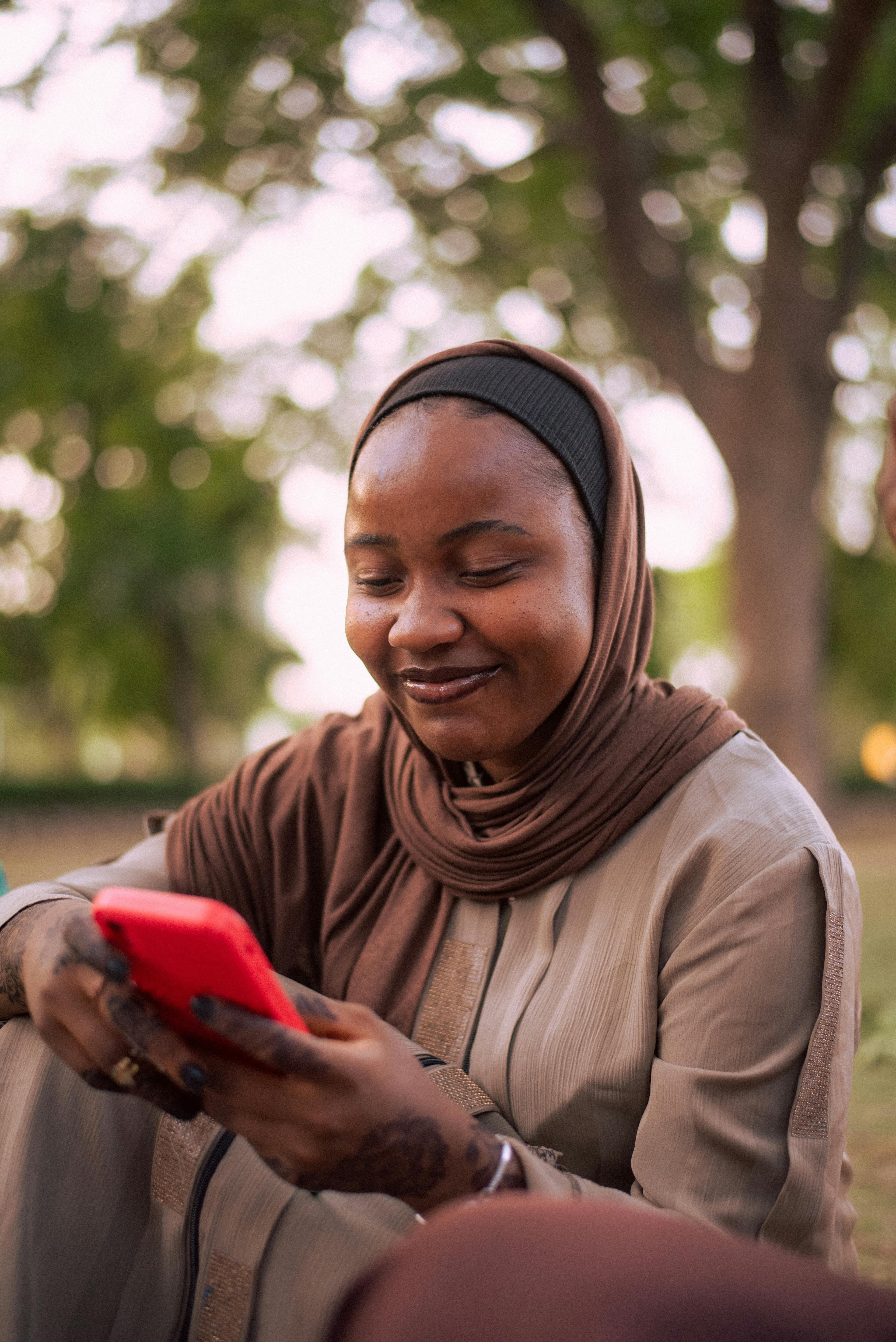 Femme en hijab marron souriante regardant son téléphone rouge, extérieur