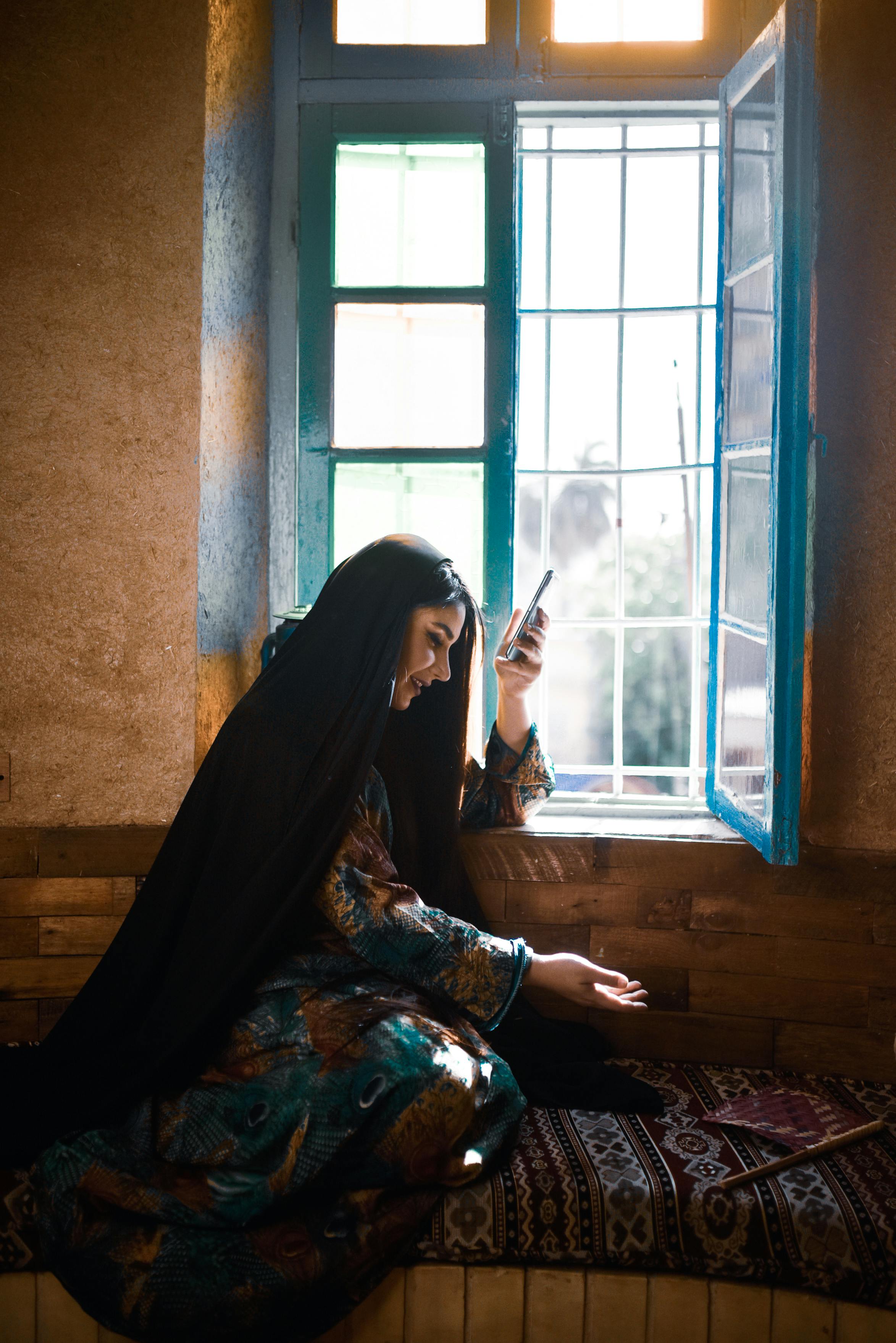 Femme afghane assise près d'une fenêtre bleue tenant son téléphone